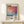 May include: Framed art print featuring abstract watercolor artwork in orange, pink, and teal tones with "Mark Rothko" text and descriptive paragraph below. Light wood frame displayed on white wooden shelf beside small wrapped gift with twine. Cream colored wall background with soft natural lighting.