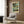 May include: Mark Rothko abstract art poster with teal, gold, and dark geometric blocks displayed in white frame on wood-paneled wall above beige armchair with geometric pillow. Modern reading nook features brass floor lamp and dark side table with open book near window with white curtains.