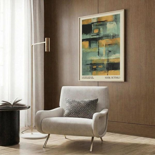 May include: Mark Rothko abstract art poster with teal, gold, and dark geometric blocks displayed in white frame on wood-paneled wall above beige armchair with geometric pillow. Modern reading nook features brass floor lamp and dark side table with open book near window with white curtains.