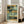 May include: Abstract art print in light wood frame leaning against beige armchair in bright living room. Artwork features geometric blocks in muted teal, sage green, golden yellow, and charcoal gray with soft brushstrokes and layered composition. Print shows artist name at bottom. Natural light streams through white-trimmed windows onto hardwood floors.