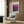 May include: A framed Mark Rothko exhibition poster with purple and magenta abstract color fields hanging on wood-paneled wall above a beige upholstered armchair with geometric patterned pillow. Modern brass floor lamp and dark side table with open book complete the contemporary living room setting with natural lighting.