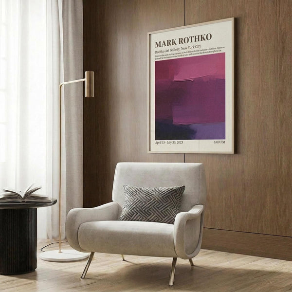 May include: A framed Mark Rothko exhibition poster with purple and magenta abstract color fields hanging on wood-paneled wall above a beige upholstered armchair with geometric patterned pillow. Modern brass floor lamp and dark side table with open book complete the contemporary living room setting with natural lighting.