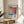 May include: Framed art print featuring Mark Rothko's name and exhibition dates 28.01-08.06.1970, with abstract watercolor-style artwork in warm orange, pink, and cool blue tones. The cream-colored poster sits on a round wooden side table next to a beige upholstered chair, with plant leaves casting shadows on white walls in natural sunlight.