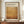 May include: Framed exhibition poster featuring Mark Rothko's name in bold black letters above warm golden-brown abstract color field artwork. The cream-colored poster shows gallery information and exhibition dates, displayed in dark wooden frame on wooden sideboard next to white ceramic vase and stacked books against cream paneled wall.