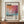 May include: Framed poster featuring abstract watercolor artwork in coral, pink, orange and turquoise tones with "Mark Rothko" text at top and exhibition details below. Displayed on wooden credenza beside white ceramic vase and stacked books against cream walls with white trim molding.