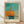 May include: Framed abstract art print leaning against white textured wall on light wood floor. Artwork features turquoise and orange geometric forms suggesting a sailboat, with mast-like vertical lines. Natural light creates diagonal shadows across the scene. Light wood frame surrounds the minimalist composition with cream matting and text below the image.