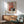 May include: Framed Mark Rothko abstract art print hanging on white wall above wooden credenza. Artwork features soft geometric rectangles in warm coral pink, orange, and deep green tones. Mid-century modern room setting with sage green upholstered armchair, round wooden side table, and white ceramic table lamp. Natural sunlight creates dramatic shadows across the minimalist interior space.