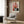 May include: Abstract art poster with orange, pink, red, and black color blocks in white frame, displayed above a cream armchair with geometric pillow. Modern minimalist room setting with wood paneled wall, brass floor lamp, and dark side table with open book.
