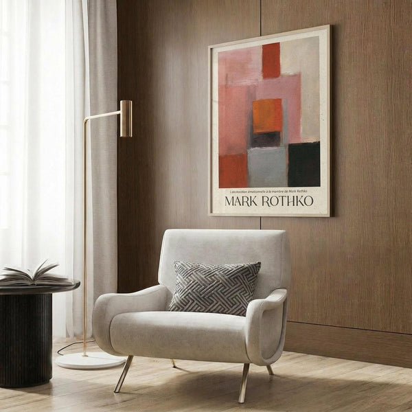 May include: Abstract art poster with orange, pink, red, and black color blocks in white frame, displayed above a cream armchair with geometric pillow. Modern minimalist room setting with wood paneled wall, brass floor lamp, and dark side table with open book.