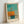 Framed abstract art print featuring a minimalist composition with turquoise sky, orange geometric ship form with dark mast details, and teal water reflection below. The artwork uses soft, muted colors in a modernist style. Displayed in a light wood frame leaning against a white wall, with visible text attribution at bottom of print.