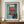 May include: Framed abstract art print displayed on wooden sideboard featuring red rectangular forms and white elements against blue background, accompanied by small white ceramic vase and stacked books in elegant room with white paneled walls.