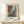 May include: Framed abstract art print with geometric color blocks in rust orange, sage green, cream, and dark tones, displayed on light wood console table against cream wall. Small wrapped gift box sits in front of frame. Natural lighting creates soft shadows.