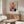 May include: Abstract art poster with geometric blocks in orange, red, pink, and dark colors on cream background, displayed above wooden sideboard in minimalist living room with white curved chair, glass coffee table, and brass floor lamp