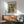 May include: Abstract art poster featuring colorful geometric squares in teal, orange, yellow, and burgundy tones displayed in wooden frame above mid-century modern credenza. Scene shows mint green upholstered chair with wooden arms, white table lamp, and round coffee table in minimalist living room with white walls and natural sunlight casting shadows.