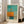 May include: Framed abstract art print leaning against white wall next to beige sofa. Artwork features turquoise sky, orange geometric ship form with mast, and teal water reflection below dark horizontal line. Light wood frame with white mat. French text at bottom reads "L'abstraction émotionnelle à la manière de Mark Rothko" and "MARK ROTHKO". Natural lighting from tall windows creates soft shadows on white tile floor.