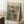 May include: Abstract art poster in light wood frame leaning against white paneled wall on textured cream rug. Artwork features geometric rectangular shapes in muted teal, coral, yellow and beige tones with soft, painterly edges and "MARK ROTHKO" text at top. Scene includes white coffee cup on wooden block side table and beige knitted throw blanket corner.