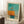 May include: Framed abstract sailboat artwork leaning against white wall corner in bright room with natural light. Turquoise sky background with orange and yellow boat forms, displayed in light wood frame. Coffee cup on wooden block and textured throw blanket visible nearby on white fluffy rug.