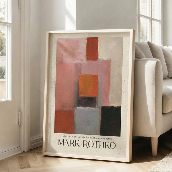 May include: Abstract art poster in light wooden frame leaning against white wall next to beige sofa. Artwork features geometric blocks in warm tones - coral pink, burnt orange, rust red, gray, and dark green rectangles arranged vertically on cream background. Text reads "Mark Rothko" at bottom. Natural lighting creates soft shadows on hardwood floor in minimalist living room setting.