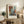 May include: Abstract geometric poster with orange, blue, and gray rectangular blocks in wooden frame, displayed on rustic wooden sideboard alongside potted fern, ceramic vase, stacked books, and beige armchair in warm neutral living room setting