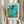 May include: Person holding framed abstract art print in light wooden frame against white curtained window. Artwork features vibrant turquoise blue background with yellow, orange, and purple geometric shapes, white dots and marks scattered throughout. Person wears cream ribbed sweater with puffy sleeves, holds dried pampas grass in other hand. Soft natural lighting creates shadows on white walls.
