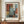 May include: Framed abstract art poster with overlapping rectangular blocks in rust orange, blue-gray, and white tones displayed on wooden credenza beside ceramic vase and books against cream paneled wall