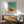 May include: Framed Mark Rothko abstract art print hanging above wooden mid-century modern credenza with white ceramic table lamp. Mint green upholstered chair with wooden arms sits nearby on light wood flooring. Artwork features orange and teal color blocks in soft, painterly style. Natural sunlight creates geometric shadows on white wall, giving the modern living room scene a warm, contemplative atmosphere.