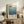 May include: Framed vintage-style art poster featuring blue gradient colors and "Mark Rothko" text, displayed on rustic wooden sideboard alongside fern plant, ceramic vase, books, and beige upholstered chair in warm-toned living room setting.