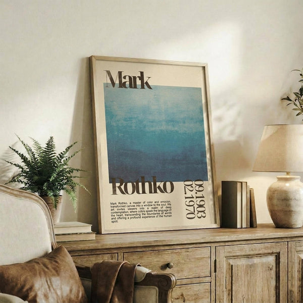 May include: Framed vintage-style art poster featuring blue gradient colors and "Mark Rothko" text, displayed on rustic wooden sideboard alongside fern plant, ceramic vase, books, and beige upholstered chair in warm-toned living room setting.