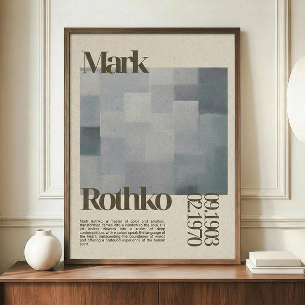 May include: Framed Mark Rothko exhibition poster with soft blue-gray abstract painting, brown text on cream background, displayed on wooden credenza beside white ceramic vase and books against beige paneled wall in modern interior setting.