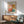 May include: Framed Mark Rothko exhibition poster hanging above a wooden sideboard with white ceramic lamp, beside a mint green mid-century armchair with wooden arms. The poster features abstract color blocks in coral, orange, teal and cream tones with black text and exhibition details. Warm sunlight creates geometric shadows across the white wall and modern furniture in this minimalist living room setting.