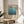 May include: Framed art print featuring Mark Rothko's name and exhibition details from 1970, with blue gradient watercolor background transitioning from light to deep blue. Print displayed on round wooden side table next to beige upholstered chair, with plant leaves visible and natural sunlight casting shadows on white wall behind.
