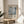 May include: Framed poster with Mark Rothko's name and exhibition details, featuring abstract geometric squares in muted blues, grays and whites. Natural wood frame sits on round wooden side table next to beige upholstered chair. Soft natural light creates shadows across cream wall. Green plant leaves visible in corner of minimalist interior setting.