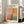 May include: Framed art poster featuring abstract color blocks in coral, orange, teal and yellow tones displayed against white wall next to beige upholstered sofa. Poster shows exhibition text for Mark Rothko at gallery with dates April 15 - July 30, 2023. Natural lighting from tall windows creates soft shadows on light wood floors.
