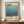 May include: Framed poster with blue gradient watercolor artwork and "Mark Rothko" typography, displayed on wooden credenza next to white ceramic vase and stacked books against cream paneled wall. The print features soft blue tones transitioning from light to deep blue with descriptive text below in brown wooden frame.