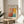 May include: Framed Mark Rothko exhibition poster displayed on light wood round side table next to beige upholstered chair. Poster features abstract colorful blocks in coral, orange, yellow, and turquoise with black text header and exhibition details. Thin gold frame catches natural sunlight streaming across neutral wall. Green plant leaves visible in corner, creating warm minimalist home decor scene.