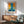 May include: Abstract art poster with vertical blue stripe through orange and yellow color fields, displayed in wooden frame above mid-century modern credenza. Room features mint green upholstered chair with wooden arms, white ceramic table lamp, and warm sunlight casting shadows on white wall.
