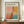 May include: Framed exhibition poster featuring Mark Rothko's abstract colorfield painting with soft blocks of coral pink, orange, yellow, red, and teal blue. The cream-colored poster displays gallery information and exhibition dates. Wooden frame sits on natural wood console beside white ceramic vase and stacked books against white paneled wall.