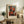 May include: Abstract art poster with red, orange, and black rectangular color blocks in wooden frame, displayed on rustic wood sideboard alongside fern plant, ceramic vase, books, and table lamp in neutral-toned living room setting with beige armchair.