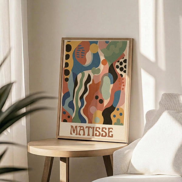 May include: Framed abstract art print in wooden frame displaying colorful organic shapes in blues, oranges, greens, and yellows with black dots and flowing forms, titled "MATISSE" at bottom. Print sits on round wooden side table next to beige upholstered chair with plant leaves visible in soft natural lighting.