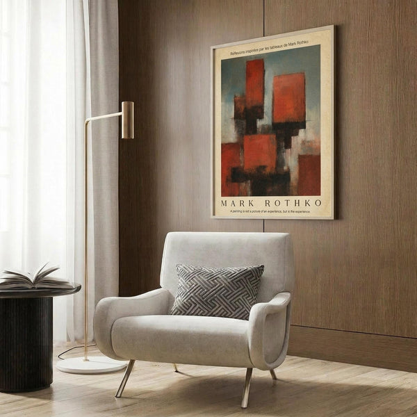 May include: Abstract art poster with red and orange rectangular forms against blue-gray background, displayed in cream frame on wood-paneled wall. Modern reading nook features beige upholstered armchair with geometric patterned pillow, brass floor lamp with white base, and dark round side table with open book. Neutral interior with curtained window and warm wood flooring.