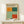 May include: Framed exhibition poster featuring Mark Rothko's abstract color field artwork with orange, teal, sage green, and coral rectangular blocks. Displayed on light wood surface beside small white gift box with twine and greenery. Natural lighting creates soft shadows on cream wall background.