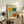 May include: Framed Mark Rothko exhibition poster with colorful geometric blocks in orange, teal, mint green, and coral red, displayed on rustic wooden sideboard alongside potted fern, ceramic vase, and books in cozy living room setting with beige chair and neutral walls