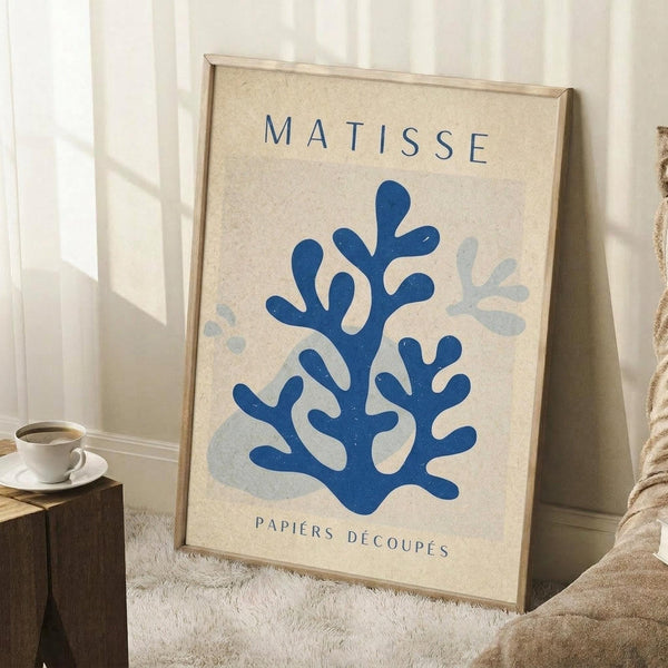 May include: Framed poster with cream background featuring blue coral-like cut-out shapes in Matisse style, displaying "MATISSE" and "PAPIERS DÉCOUPÉS" text. Light wood frame leaning against white wall corner near wooden side table with white coffee cup and beige throw blanket on textured rug.