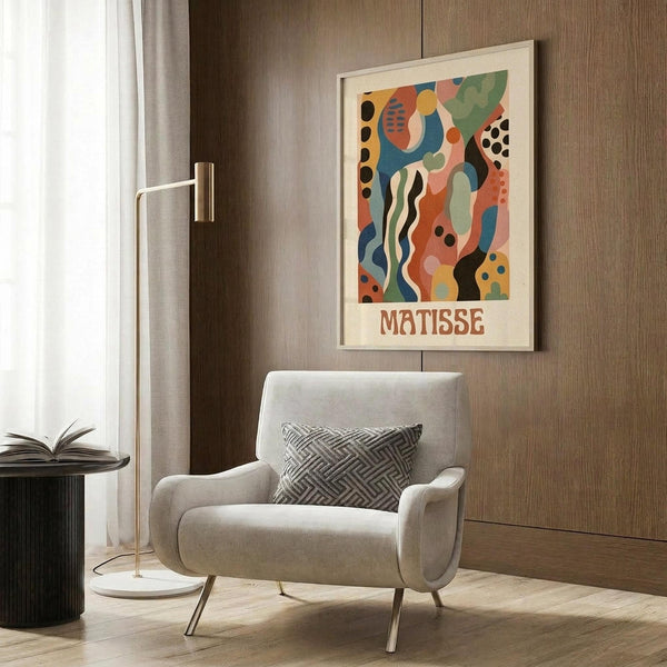 May include: Colorful abstract art poster with organic shapes in coral, blue, green, and orange tones, featuring "MATISSE" text below the artwork. Displayed in light wood frame on wood-paneled wall above cream armchair with geometric pillow. Modern room setting with brass floor lamp and side table.