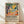 May include: Framed poster with colorful tropical scene featuring yellow sun, green palm fronds, orange citrus fruits, and coral pink brushstrokes in Matisse-inspired style. Black text below reads "Matisse: A Master of Color and Form" with exhibition description. Natural wood frame sits on light wooden floor against white wall with soft shadows.