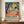 May include: Framed exhibition poster featuring colorful abstract artwork with orange sun, blue trees, green foliage, and coral ground in Matisse-inspired style. Black text below describes art exhibition. Poster displayed on wooden credenza beside white ceramic vase and books in elegant room with white paneled walls.