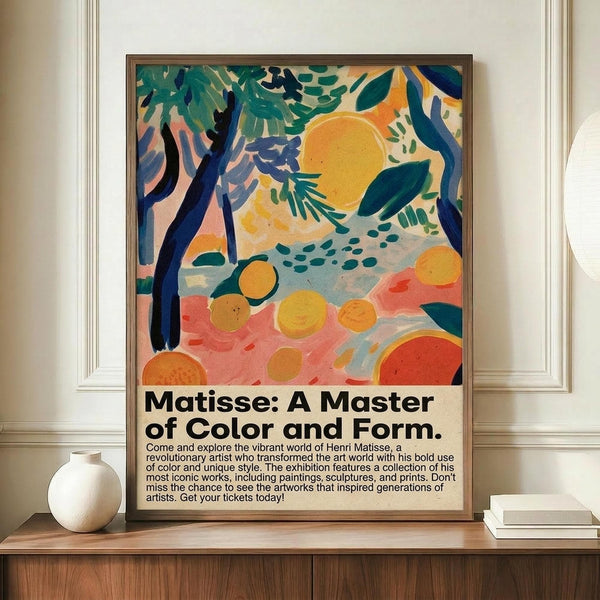 May include: Framed exhibition poster featuring colorful abstract artwork with orange sun, blue trees, green foliage, and coral ground in Matisse-inspired style. Black text below describes art exhibition. Poster displayed on wooden credenza beside white ceramic vase and books in elegant room with white paneled walls.