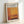 May include: Framed exhibition poster for Mark Rothko at Rothko Art Gallery, New York City, featuring abstract orange and brown color field artwork. Light wood frame sits propped against white wall with natural lighting creating soft shadows. Poster shows exhibition dates April 15 - July 30, 2023 at 6:00 PM with descriptive text about Rothko's transcendental color and emotion work.