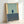 May include: Framed poster print in natural wood frame leaning against white wall. Abstract minimalist artwork features geometric color blocks - pale mint green upper section with yellow vertical strip on left, small dark blue rectangle in center, and darker blue-gray lower section. Artist name "MARK ROTHKO" printed in small serif text at bottom right. Modern contemporary art style with clean lines and muted color palette.