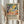 May include: Person holding framed art print featuring abstract landscape with yellow sun, green foliage, orange fruits, and purple tree branches in Matisse-inspired style. Light wood frame against neutral curtained background with dried pampas grass visible. Cream-colored sweater with gathered sleeves.